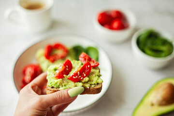 Wholesome healthy breakfast - a woman's hand holding a vegan avocado sandwich on wholewheat bread, topped with fresh cherry tomatoes — a clean, nourishing choice for mindful eating and balanced living