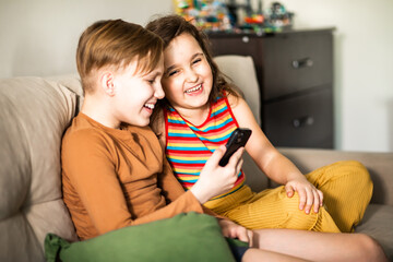 Cute little girl and boy teenager using smart phone sitting on sofa in living room at home together.