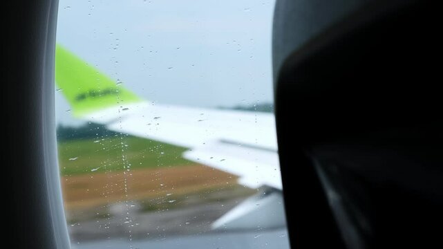 The plane picks up speed on the runway and takes off, lifting off from the ground. Raindrops are running down the glass. View the porthole to the wing of moving passenger airplane. HQ 4K footage.