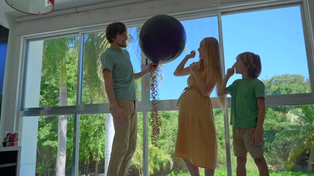 Happy family celebrating indoor gender reveal. Pregnant mom pops balloon with pink confetti, revealing it's a girl. Dad and big brother hug and cheer.