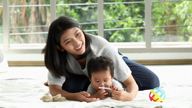 Young mother lying on white blanket playfully bonding with curious baby holding teether toy on bright indoor floor near large glass window