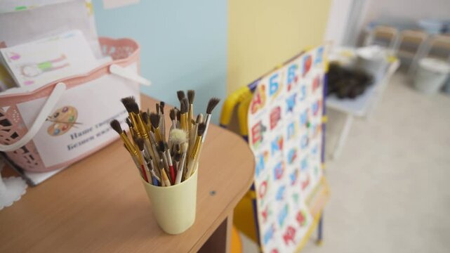 Paintbrushes stand in plastic cup on wooden desk in colorful classroom. Art supplies rest beside cheerful alphabet chart on nearby wall