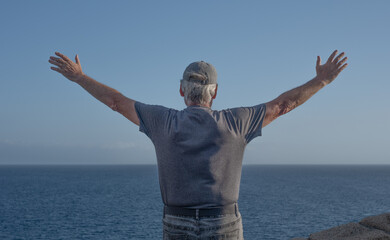 Rear view of senior man along the coast looking at the horizon over sea with outstretched arms,...