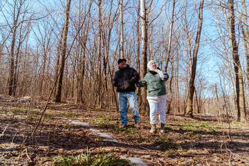 Fototapeta premium An elderly couple, a man and a woman, walk through a spring forest, enjoying the beauty of nature. Sunlight filters through fresh green leaves, creating a serene and romantic atmosphere