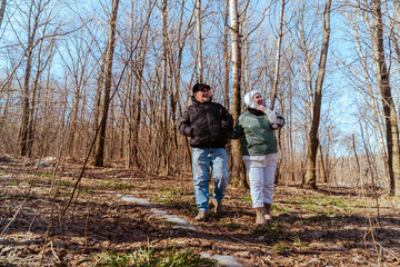 An elderly couple, a man and a woman, walk through a spring forest, enjoying the beauty of nature. Sunlight filters through fresh green leaves, creating a serene and romantic atmosphere