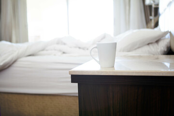 white cup on the bedside table near empty white bed sheets, seen through large window.