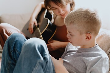 Mother and son playing guitar together, family love. The child wears a hearing aid, highlighting...