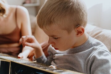 Mother and son playing guitar together, family love. The child wears a hearing aid, highlighting...