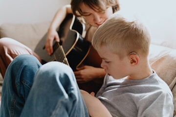 Mother and son playing guitar together, family love. The child wears a hearing aid, highlighting...