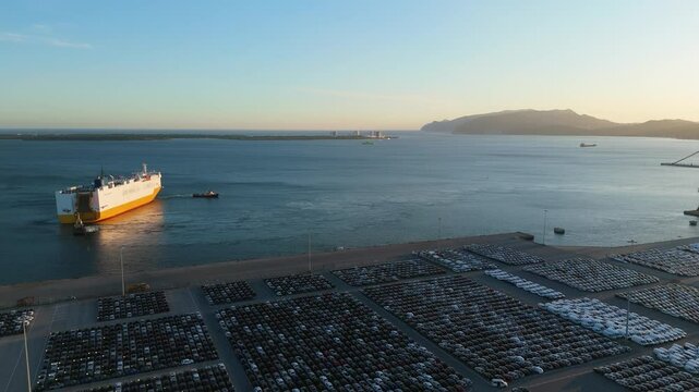 Ro-Ro Cargo Ship with Tugboats Arriving and Rows of New Cars on Large Full Parking Lot at Sunset in Port. Automotive Industry. Setubal, Portugal. Aerial View. Moving Forward