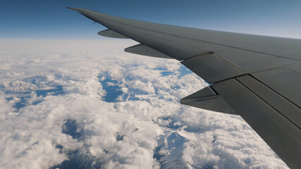 Fototapeta premium Flying on plane, view from window on wing. Flying over high snow capped mountains range and white clouds.