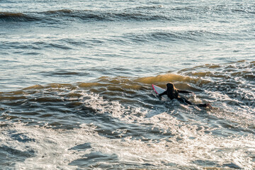 A young female surfer in a black wetsuit paddles through the ocean waves off the coast of Lima, Peru