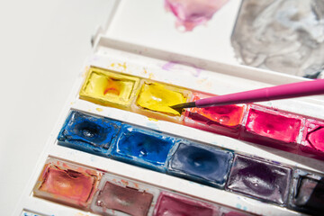 Bright watercolor palette with pink brush and vibrant mix of yellow, blue, and red paints.