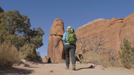 Mature active overweight female hiker with backpack hiking on hilly tourist footpath among orange buttes rock formations. Arches national park Utah USA on hot sunny day. Slow motion, back view