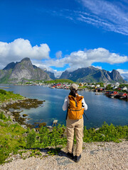 A traveler stands by the serene waters of a Norwegian fjord, surrounded by stunning mountains and charming fishing villages. The bright sky adds vibrancy to this enchanting location. Lofoten Norway