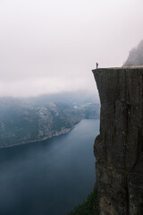 A solitary figure stands at the edge of a towering cliff, gazing out over Preikestolen Norway dramatic fjords. Dense fog envelops the mountains, enhancing the sense of adventure and awe.