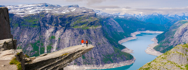 Two hikers stand on a dramatic cliff edge, overlooking Trolltunga, Norway majestic fjords and lush valleys. The sky is partly cloudy, creating a perfect backdrop for this awe-inspiring landscape.