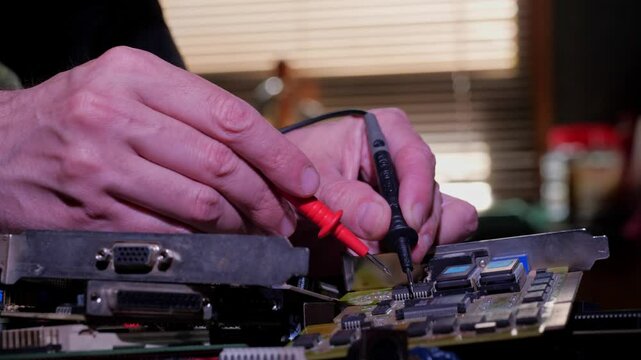 A close-up shot of a hand using a soldering iron to join wires to a circuit board. Solder fumes are visible, and various electronic components and tools are blurred in the background