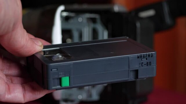 A close-up shot of a hand inserting an S-VHS video cassette into the side loading mechanism of a vintage video camera. The black cassette and the camera's textured body are clearly visible