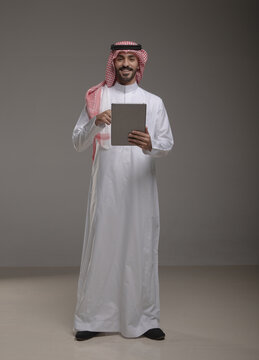 Saudi Arabian man wearing traditional thobe and shemagh using a tablet device, standing in studio with a gray background, showing screen, perfect for technology, business or communication themes.