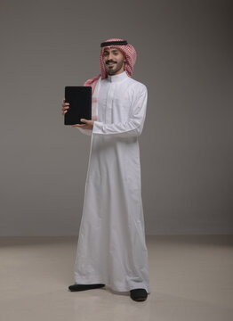 Saudi Arabian man wearing traditional thobe and shemagh using a tablet device, standing in studio with a gray background, showing screen, perfect for technology, business or communication themes.