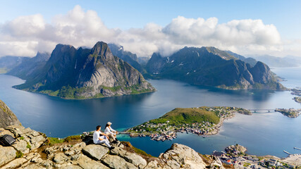 Dramatic mountains rise above pristine waters as two adventurers relax at a mountaintop in Lofoten Norway. The vibrant green of the landscape contrasts beautifully with the blue fjords below.