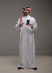 Saudi Arabian man wearing traditional thobe and shemagh using a smartphone, making a call and...