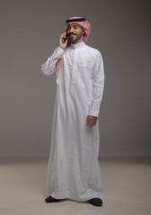 Saudi Arabian man wearing traditional thobe and shemagh using a smartphone, making a call and...