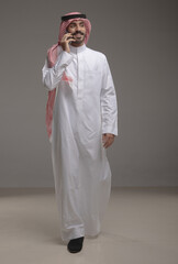 Saudi Arabian man wearing traditional thobe and shemagh using a smartphone, making a call and...