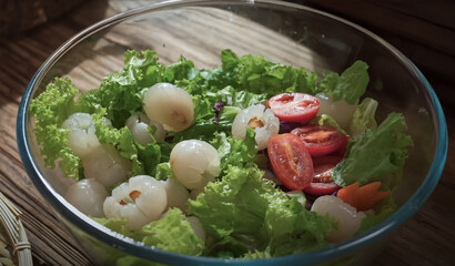 Fresh salad bowl with peeled lychees, cherry tomatoes, and leafy greens, served in a glass bowl on a wooden table with natural daylight

