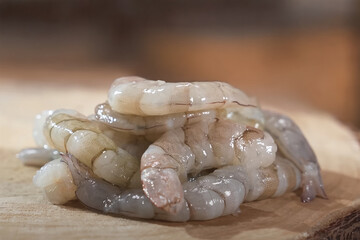 Raw shrimp without shell stacked on a wooden cutting board, showcasing fresh, glossy texture and translucent surface under soft indoor lighting

