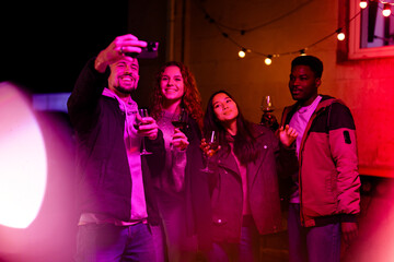 Friends enjoy a lively evening gathering while taking a selfie with drinks in a warmly lit outdoor...