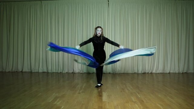 A pantomime scene, an actress juggles a poi, a long piece of cloth, on stage, making circular movements. The stage is illuminated by multi-colored iridescent lights.