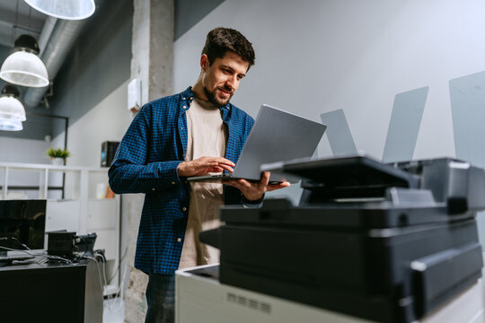 Man using laptop in modern office space while standing next to printer during working hours