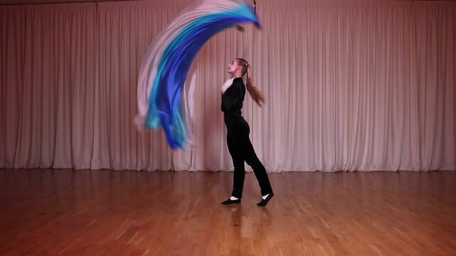 A pantomime scene, an actress juggles a poi, a long piece of cloth, on stage, making circular movements. The stage is illuminated by multi-colored iridescent lights.
