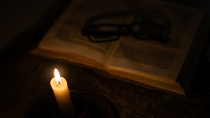 A lit candle warmly illuminates the darkness, with an open book and out-of-focus glasses in the...