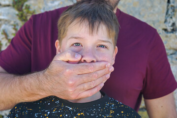 Adult hand covering young boys mouth, boy gazing at camera with distressed expression, concept of...