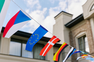 A colorful string of international flags is seen fluttering against the background of a bright building with windows. The flags are hung on a line and feature symbols of various countries, including t