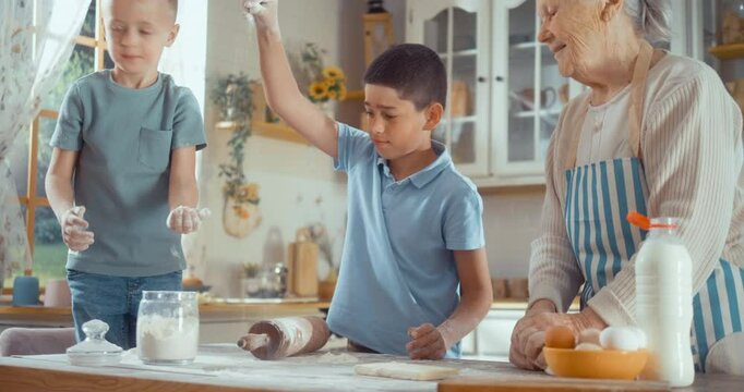Joyful 90s grandmother and grandchildren are kneading dough together in the kitchen. Children play with flour while making a pie with their grandma at home