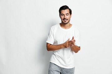 Casual smiling man wearing a white t-shirt gestures with hands while standing on a gray background....