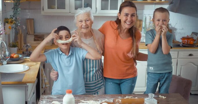 Multi-generational happy family with children enjoys cooking together in the kitchen. Portrait of 90s grandmother, adult daughter, and two children smiling at the camera.