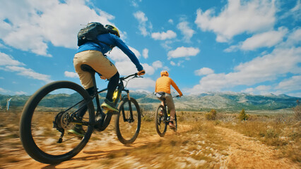Naklejka premium Two cyclists pedal down a dirt trail, surrounded by a picturesque mountain range. A close-up shot highlights their speed and the exhilaration of off-road biking.