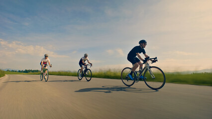 Three helmeted cyclists pedal down a scenic road, with a close-up shot highlighting their speed and focus. The lead rider, a man in a black shirt, is followed by two others in white and black.