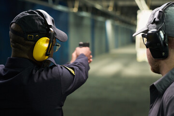 Person practicing shooting at indoor shooting range with handgun while wearing ear and eye protection. Indoor environment with professional equipment visible in background