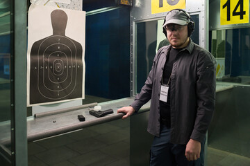 Man practicing shooting at indoor firing range wearing protective gear and serious expression while standing near firearm and target stand