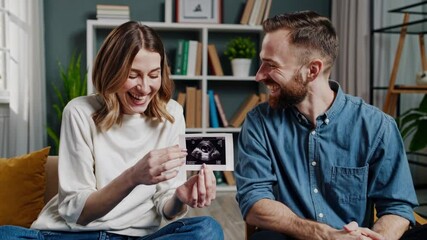 Future parents sitting on a cozy sofa, joyfully holding an ultrasound picture of their baby, sharing laughter and embracing the happiness of their pregnancy journey together - Powered by Adobe