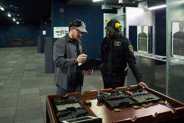 Two officers discussing tactical training while reviewing notes at indoor shooting range with various firearms and equipment visible on table