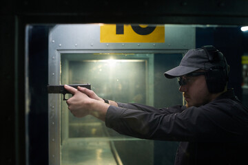 Man wearing cap and safety glasses focusing on target while standing in indoor gun range practicing shooting techniques © pressmaster