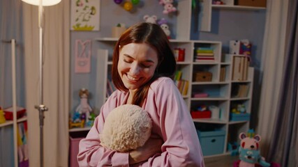 Young babysitter is enjoying her time in a colorful playroom, hugging a teddy bear and smiling with closed eyes, surrounded by toys and children's books