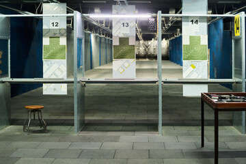 Indoor shooting range featuring several target practice stands with paper targets hanging at varying distances. Metal frames and concrete floor contribute to the industrial feel